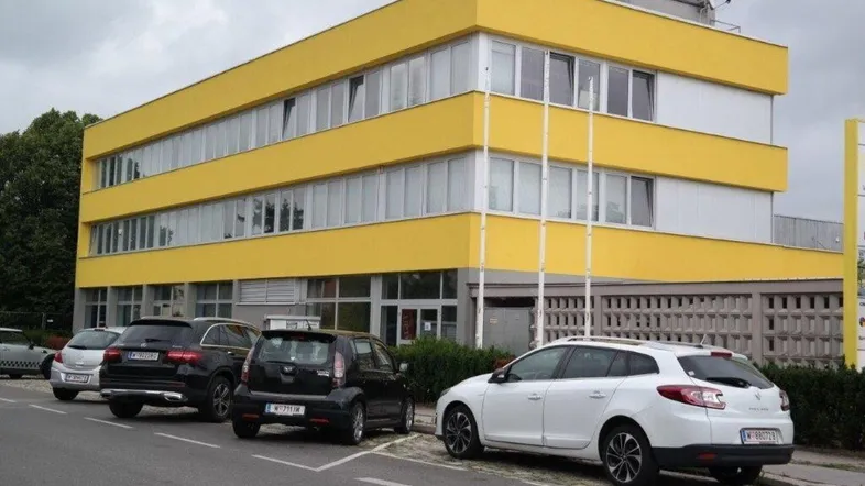 Modernes Bürogebäude mit gelber Fassade und Parkplätzen davor unter bewölktem Himmel.