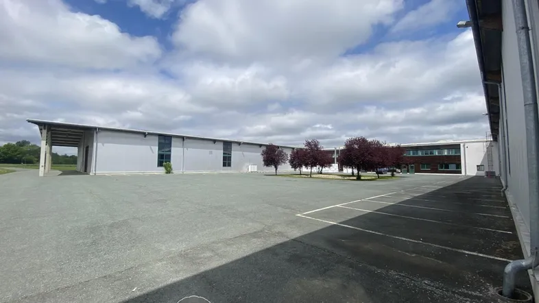 Großer asphaltierter Vorplatz vor einem modernen Industriegebäude mit weitem Himmel und vereinzelten Bäumen.