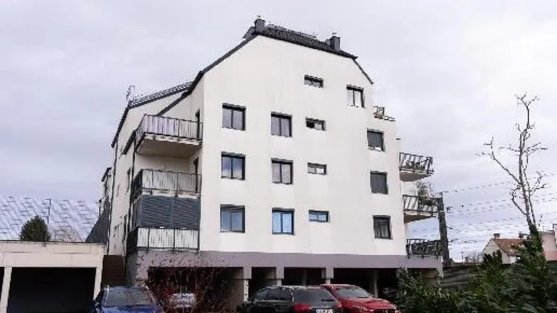 Weißes Mehrfamilienhaus mit mehreren Balkonen und Parkplätzen im Freien unter bewölktem Himmel.