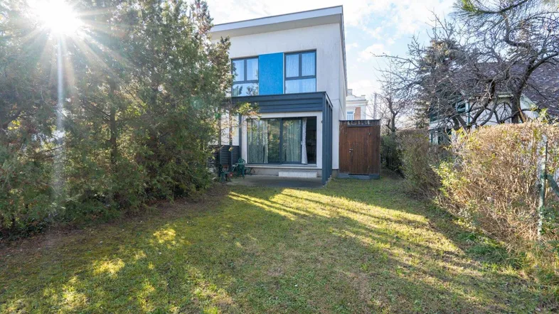 Modernes Haus mit Garten und Terrasse, umgeben von Bäumen unter strahlendem Sonnenschein.