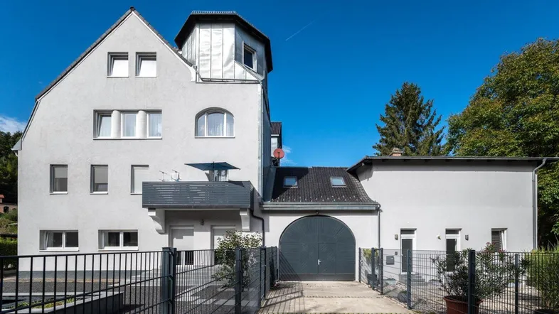 Graues Mehrfamilienhaus mit Balkon, Turmzimmer und Garageneinfahrt unter blauem Himmel.