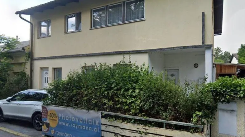 Gelbe Hausfassade mit Fenstern, umgeben von Büschen und einem Zaun an der Straße.