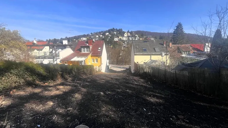 Leeres Grundstück mit Blick auf umliegende Häuser und Hügellandschaft unter blauem Himmel.