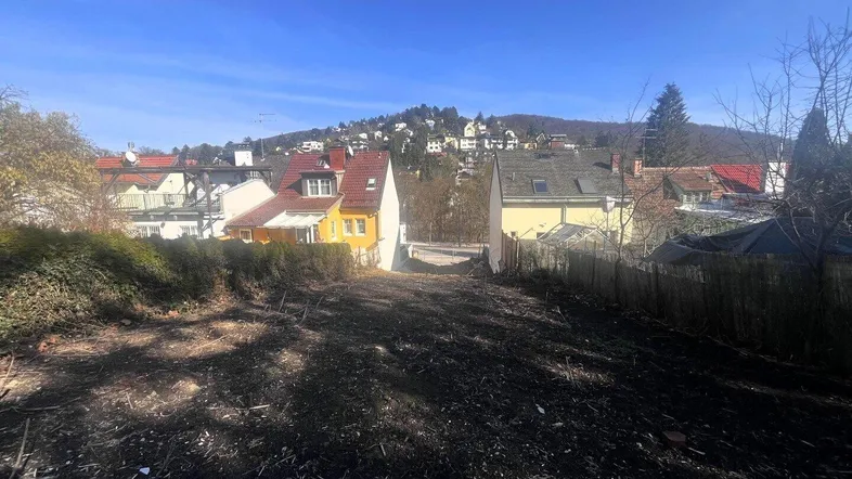 Leeres Grundstück mit Blick auf umliegende Häuser und bewaldete Hügel unter blauem Himmel.