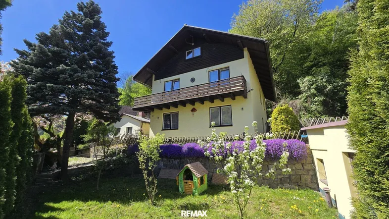 Hausfassade mit Balkon und Gartenbereich, umgeben von viel Grün und blühenden Pflanzen.