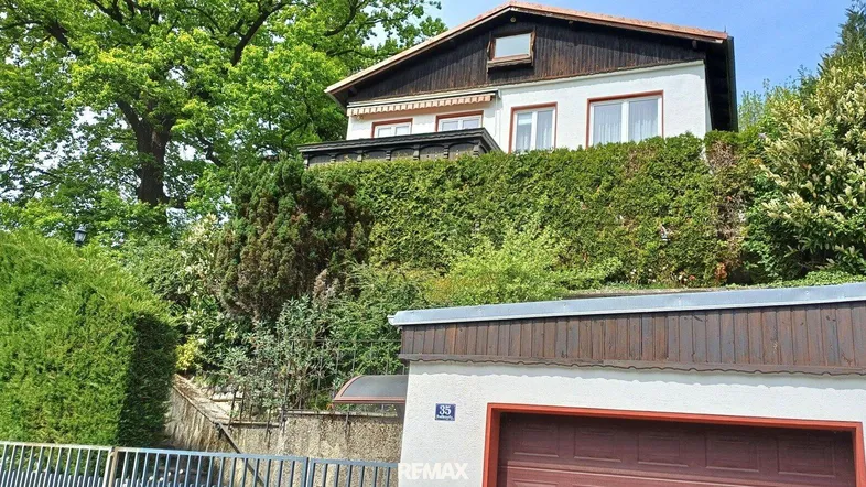 Haus auf einem Hügel mit viel Grün und einer Garage im Vordergrund, umgeben von Bäumen.