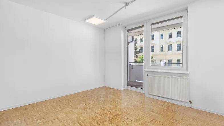 Helles, leeres Zimmer mit Parkettboden, weißen Wänden und Zugang zu einem Balkon.