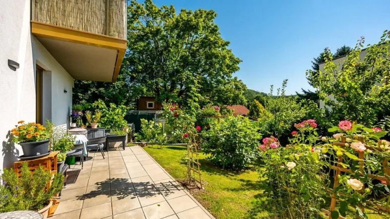 Helle Terrasse mit Fliesenboden und üppigem Garten mit Rosen und Bäumen.