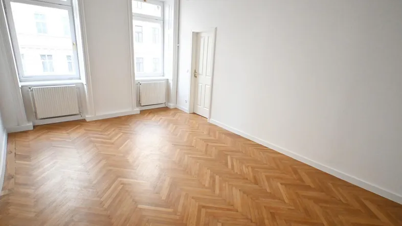 Helles Zimmer mit Fischgrätparkettboden und großen Fenstern, ideal als Wohn- oder Schlafzimmer.