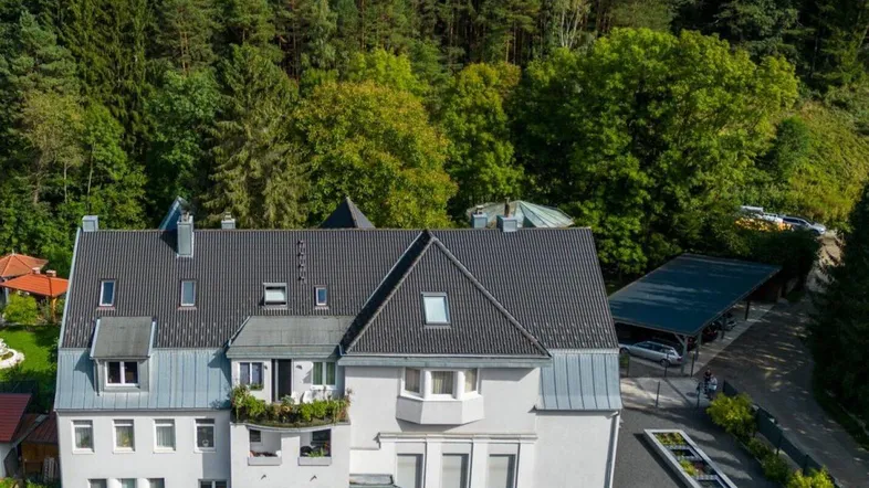 Luftaufnahme eines Mehrfamilienhauses mit dunklem Dach, umgeben von üppigem Grün und Wald.