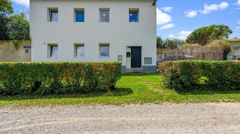 Weißes Einfamilienhaus mit gepflegter Hecke und Kiesweg im Vorgarten unter blauem Himmel.