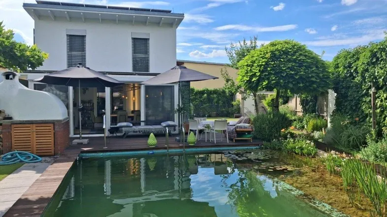 Idyllischer Gartenbereich mit Naturpool, Holzterrasse und Außenofen, umgeben von viel Grün.