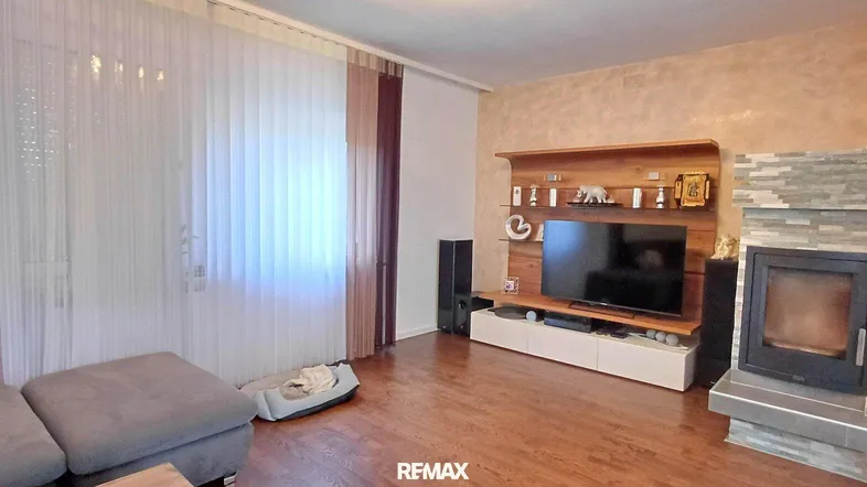 Gemütliches Wohnzimmer mit Holzboden, Kamin und modernem TV-Möbel. Helle Vorhänge lassen viel Licht herein.