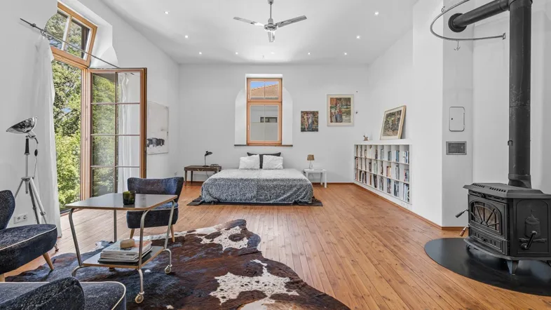 Geräumiges Schlafzimmer mit Holzboden, Kaminofen und Zugang zu einem Balkon mit Blick ins Grüne.
