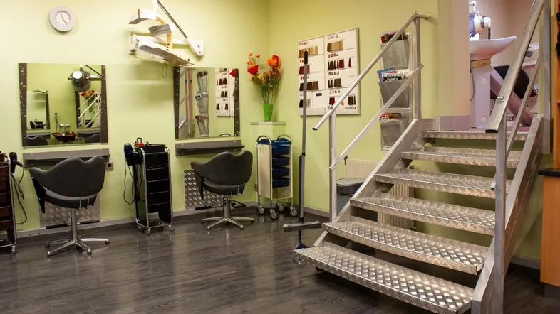 Heller Friseursalon mit mehreren Styling-Stationen, dunklem Boden und einer markanten Metalltreppe.
