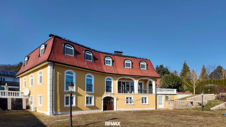 Gelbe Villa mit rotem Mansarddach und gepflegtem Vorgarten unter blauem Himmel.