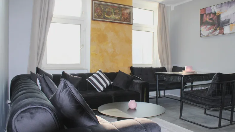 Helles Wohnzimmer mit dunklem Ecksofa, Couchtisch und einem kleinen Essbereich am Fenster.