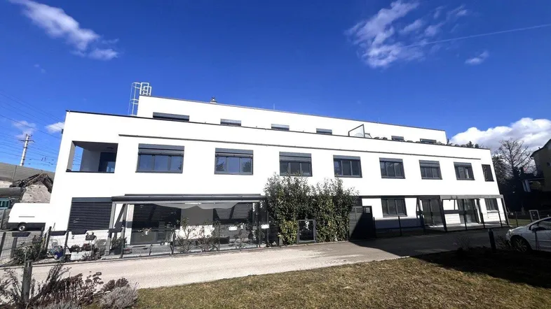 Modernes weißes Gebäude mit dunklen Fensterrahmen und klarem Himmel, umgeben von Grünflächen.