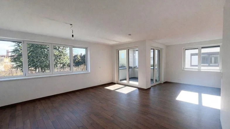 Helles Wohnzimmer mit dunklem Parkettboden, großen Fenstern und Zugang zu einem Balkon.