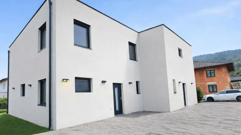 Modernes Einfamilienhaus mit weißer Fassade, dunklen Fenstern und gepflasterter Auffahrt.