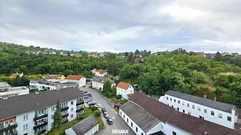 Weitläufiger Blick über eine Stadt mit grünen Hügeln und Wohngebäuden unter bewölktem Himmel.