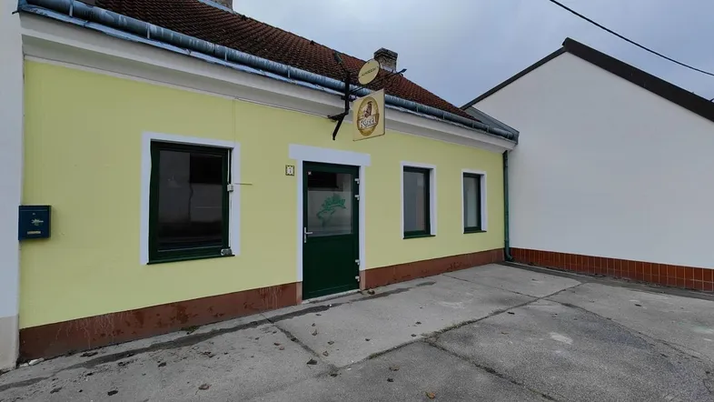 Gelbe Fassade eines älteren Gebäudes mit grüner Eingangstür und Fenstern, umgeben von einem gepflasterten Bereich.