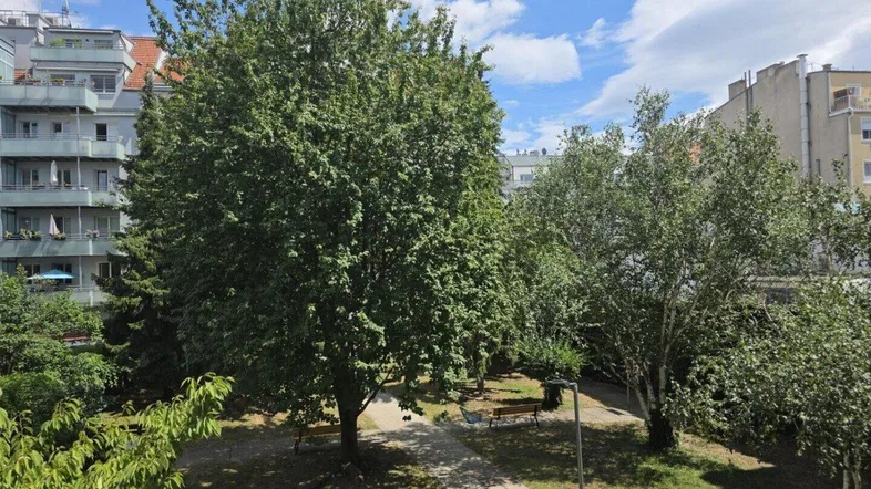 Grüner Innenhof mit Bäumen und Sitzbänken, umgeben von Wohngebäuden unter blauem Himmel.