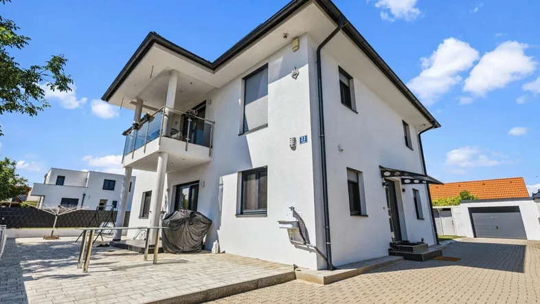 Modernes Einfamilienhaus mit weißer Fassade, Balkon und gepflastertem Vorplatz unter blauem Himmel.