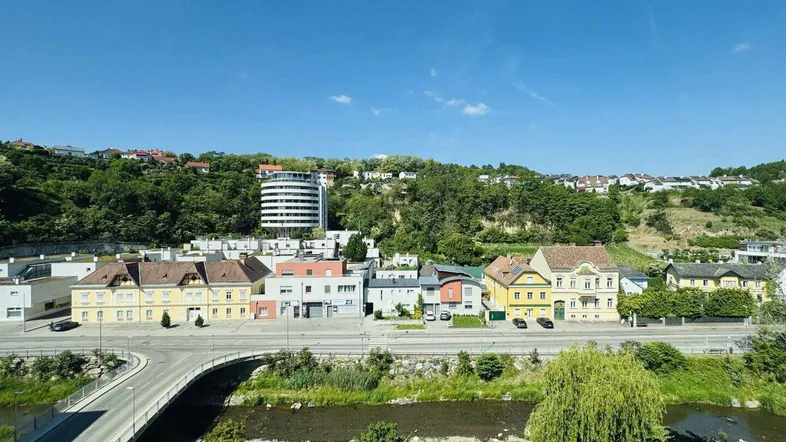 Panoramablick auf eine Stadtlandschaft mit Fluss, Brücke und bewaldeten Hügeln unter blauem Himmel.