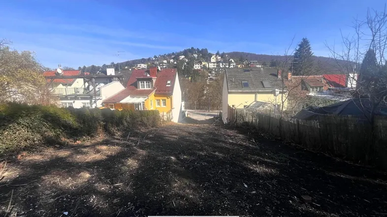 Unbebautes Grundstück mit Blick auf umliegende Häuser und eine bewaldete Hügellandschaft unter blauem Himmel.
