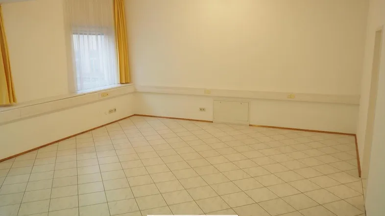 Geräumiger Büroraum mit hellen Fliesen, weißen Wänden und einem Fenster mit gelben Vorhängen.