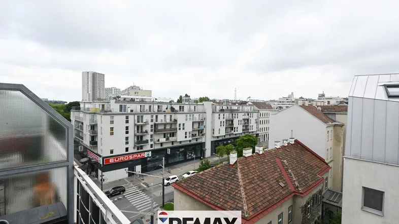 Blick über die Straße auf umliegende Gebäude und einen Supermarkt unter bewölktem Himmel.