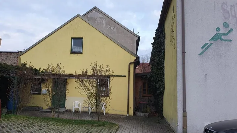 Gelbes Gebäude mit schlichter Fassade und gepflastertem Vorplatz, umgeben von spärlicher Vegetation.