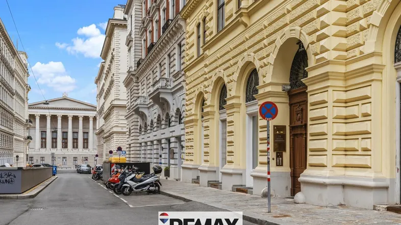 Historische Gebäudezeile in einer belebten Stadtstraße mit klassischer Architektur und geparkten Fahrzeugen.
