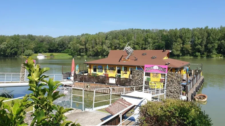 Weitläufige Ansicht des schwimmenden Gebäudes auf dem Fluss, umgeben von üppiger Natur und weiteren Booten.