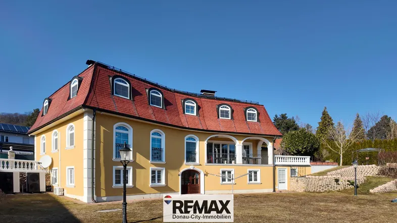 Repräsentative Villa mit gelber Fassade, rotem Mansarddach und gepflegtem Vorgarten unter blauem Himmel.