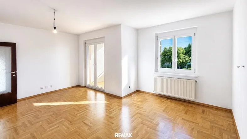 Helles Zimmer mit Fischgrätparkettboden, weißen Wänden und Zugang zu einem Balkon.