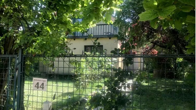 Hausfassade hinter einem grünen Zaun, umgeben von üppiger Vegetation und Bäumen.