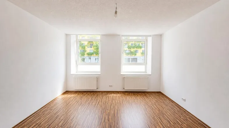 Heller, leerer Raum mit Laminatboden und zwei Fenstern, die Tageslicht hereinlassen.