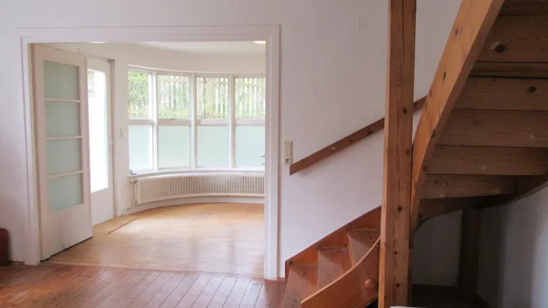 Heller Eingangsbereich mit Holztreppe und Zugang zu einem Zimmer mit Erkerfenstern.