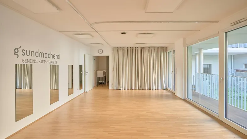 Großer, heller Raum mit Holzfußboden, Wandspiegeln und großen Fenstern, ideal für Kurse oder Therapien.