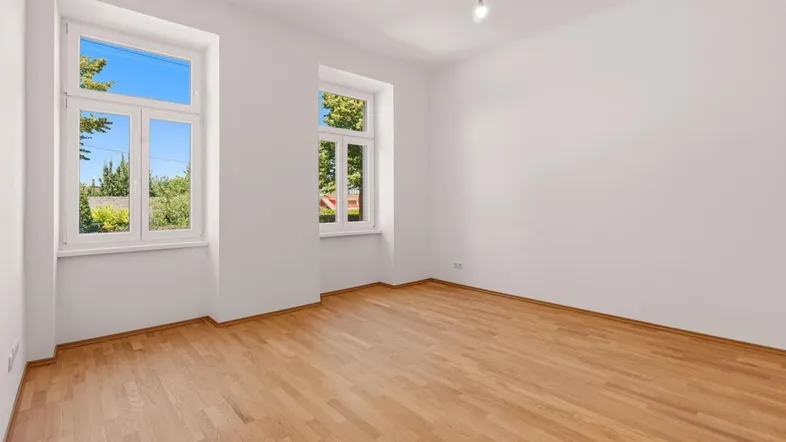 Helles Zimmer mit Parkettboden und Blick ins Grüne durch zwei große Fenster.