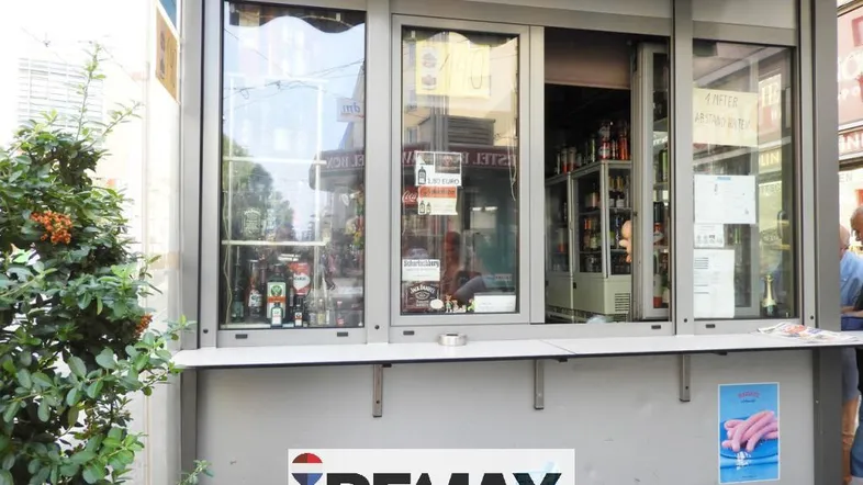 Außenansicht eines kleinen Kiosks mit Verkaufsfenstern und Werbeschildern an einer belebten Straße.