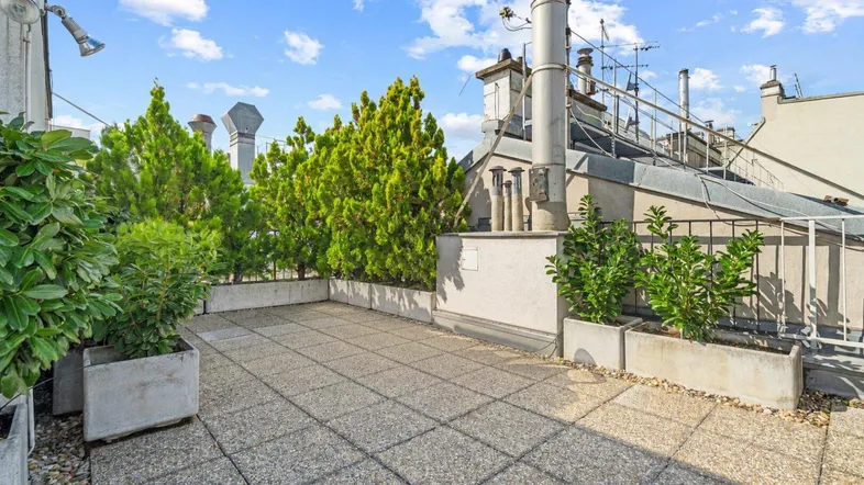 Geräumige Dachterrasse mit Steinplattenboden und grünen Pflanzen in Betonkübeln unter blauem Himmel.