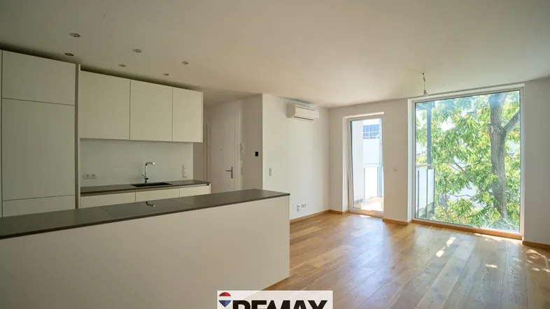 Heller offener Wohnbereich mit integrierter Küche, Parkettboden und Zugang zum Balkon.