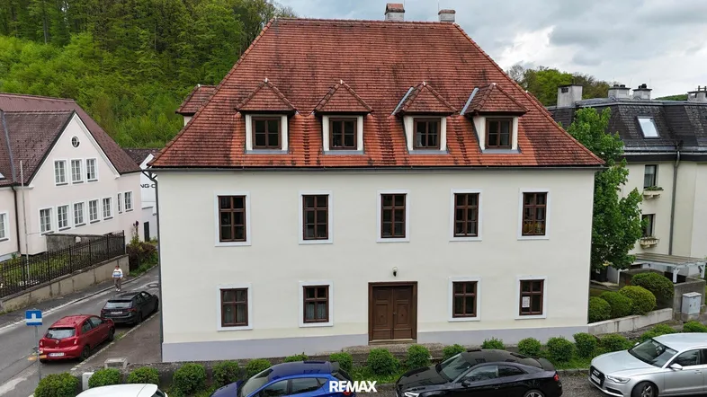 Historisches Gebäude mit heller Fassade, rotem Ziegeldach und Gaubenfenstern, umgeben von Grünflächen.