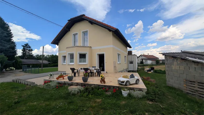 Gelbes Einfamilienhaus mit kleiner Holzterrasse und Gartenbereich unter blauem Himmel.
