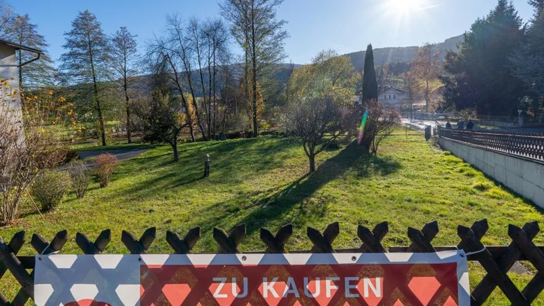 Grüne Rasenfläche mit Bäumen und einem Zaun, im Hintergrund sind Hügel und Häuser sichtbar.