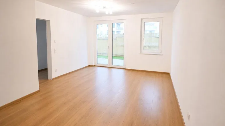 Helles, leeres Zimmer mit Laminatboden und Zugang zu einem kleinen Gartenbereich.