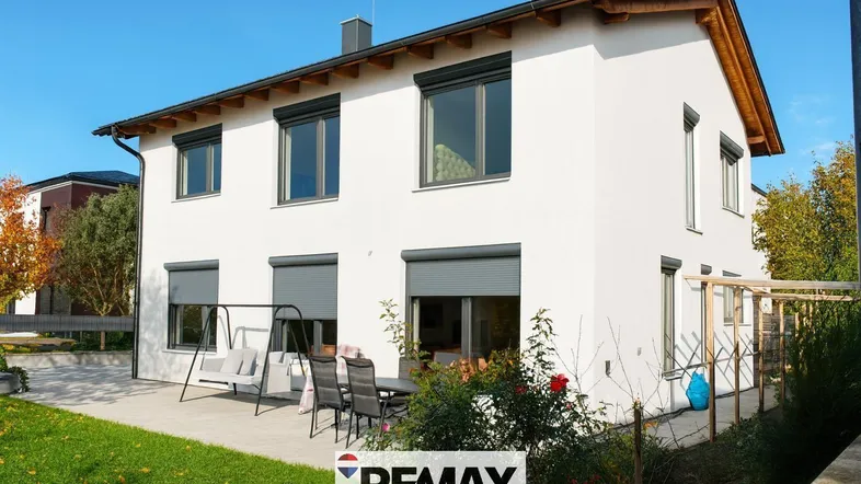 Moderne Hausfassade mit weißem Putz, Rollläden und einer möblierten Terrasse im Garten.